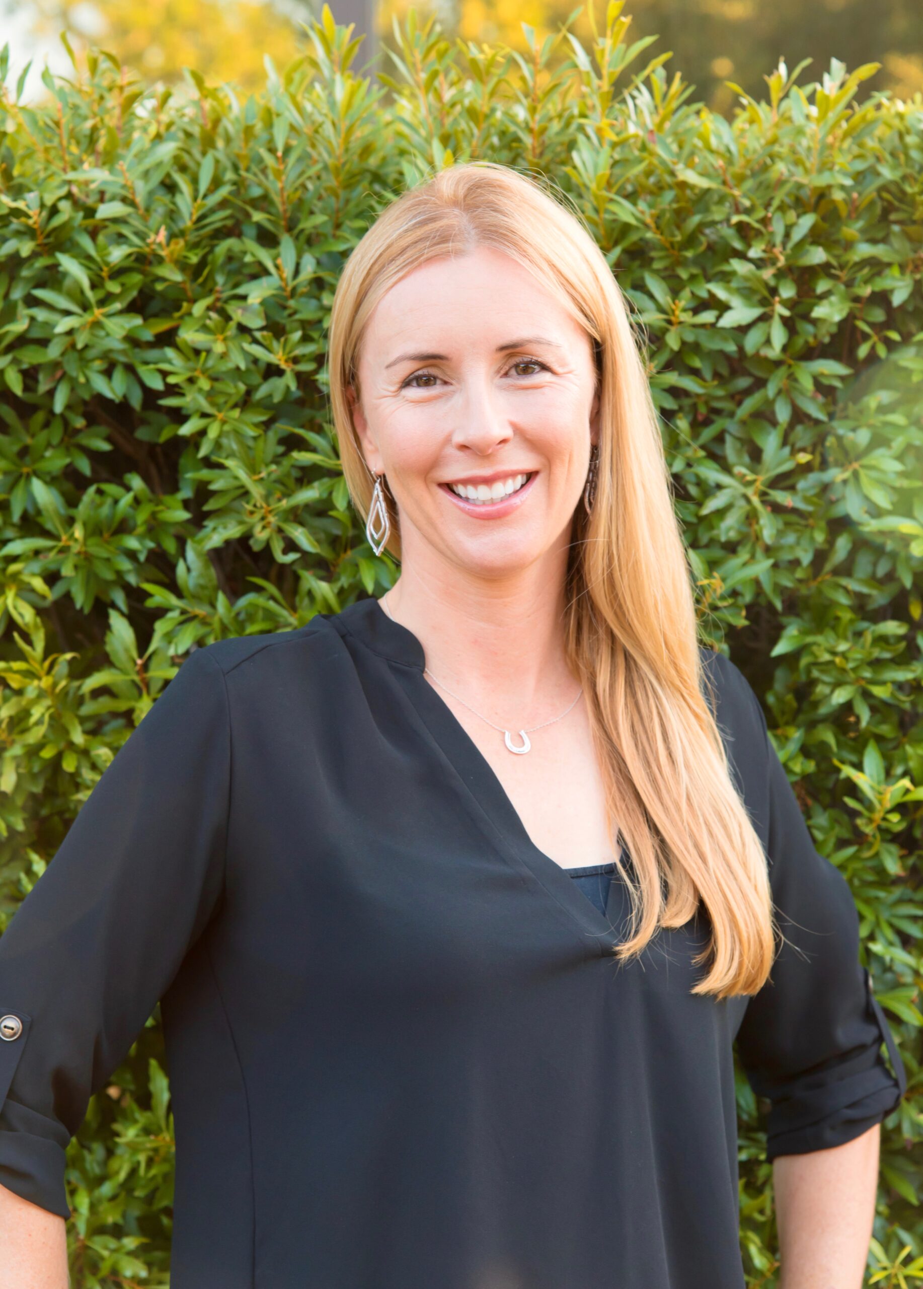 A woman standing in front of bushes smiling for the camera.