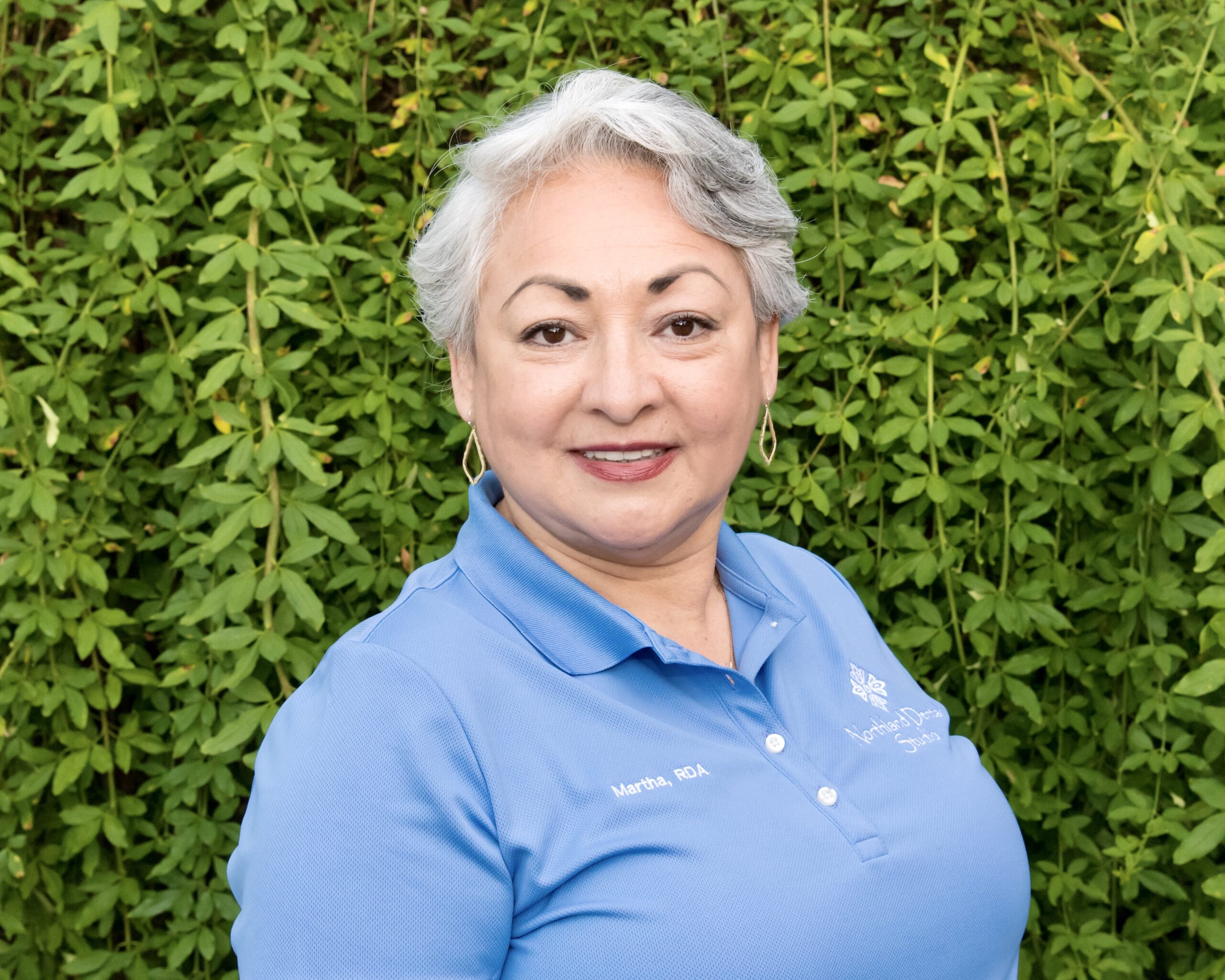 A woman standing in front of a wall with bushes.