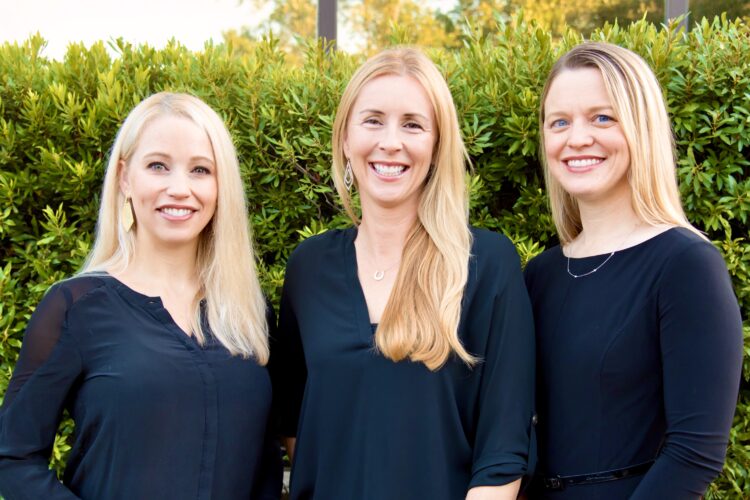 Three women standing next to each other in front of a bush.