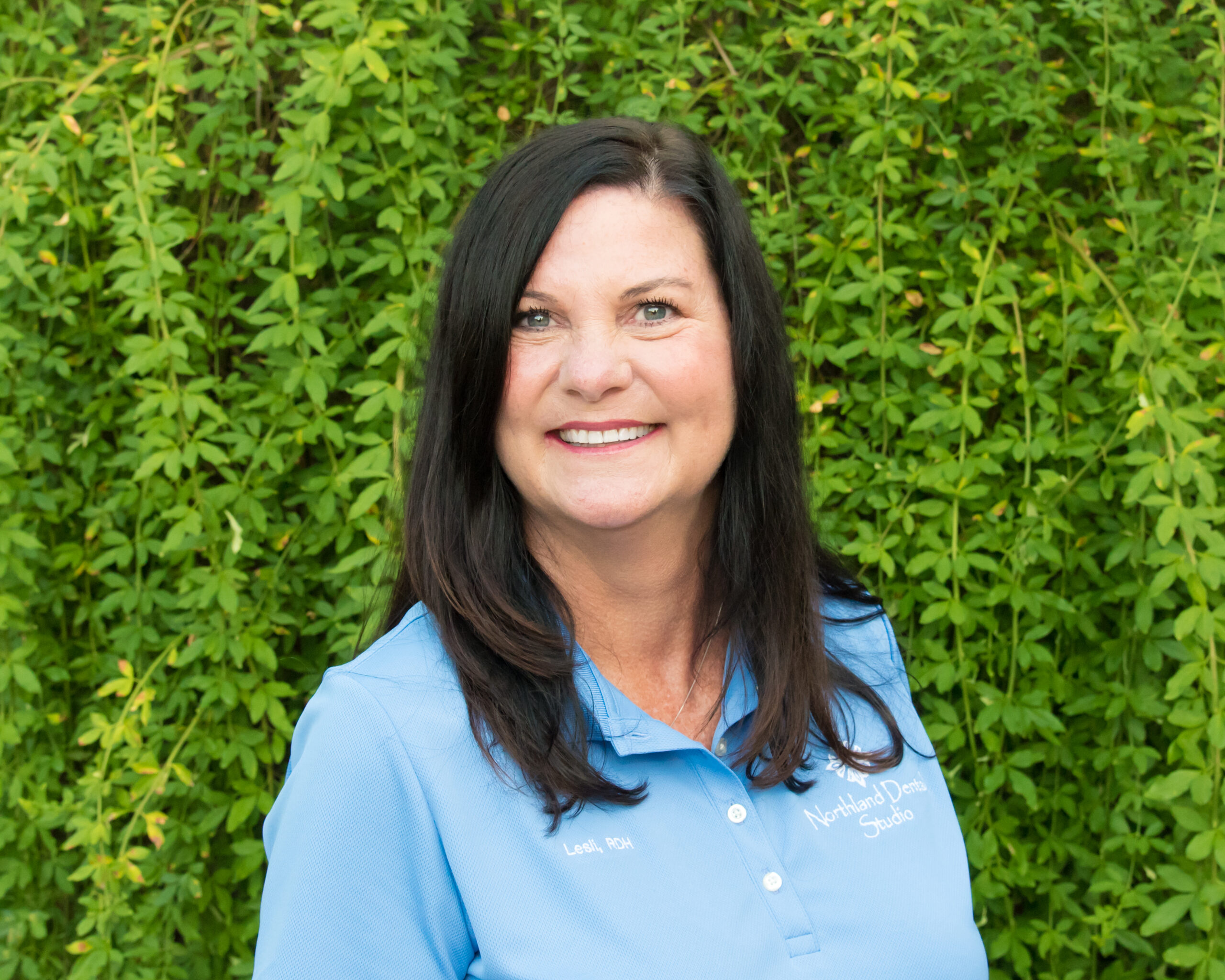 A woman standing in front of a wall with green bushes.